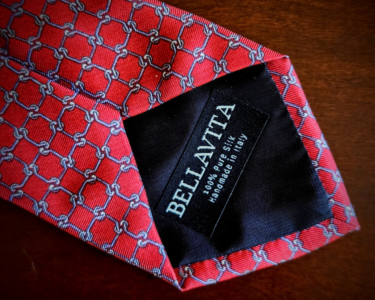 A close-up of a red necktie with a gray chain-link pattern, showing a black fabric brand label that reads BELLAVITA.
