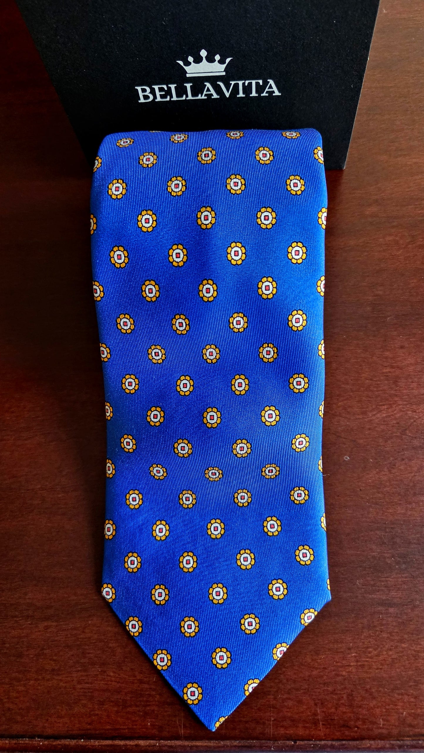 A blue silk necktie with a repeating gold and white geometric pattern, displayed flat on a dark wooden surface with a black Bellavita box behind it.