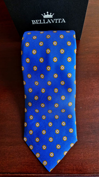 A blue silk necktie with a repeating gold and white geometric pattern, displayed flat on a dark wooden surface with a black Bellavita box behind it.