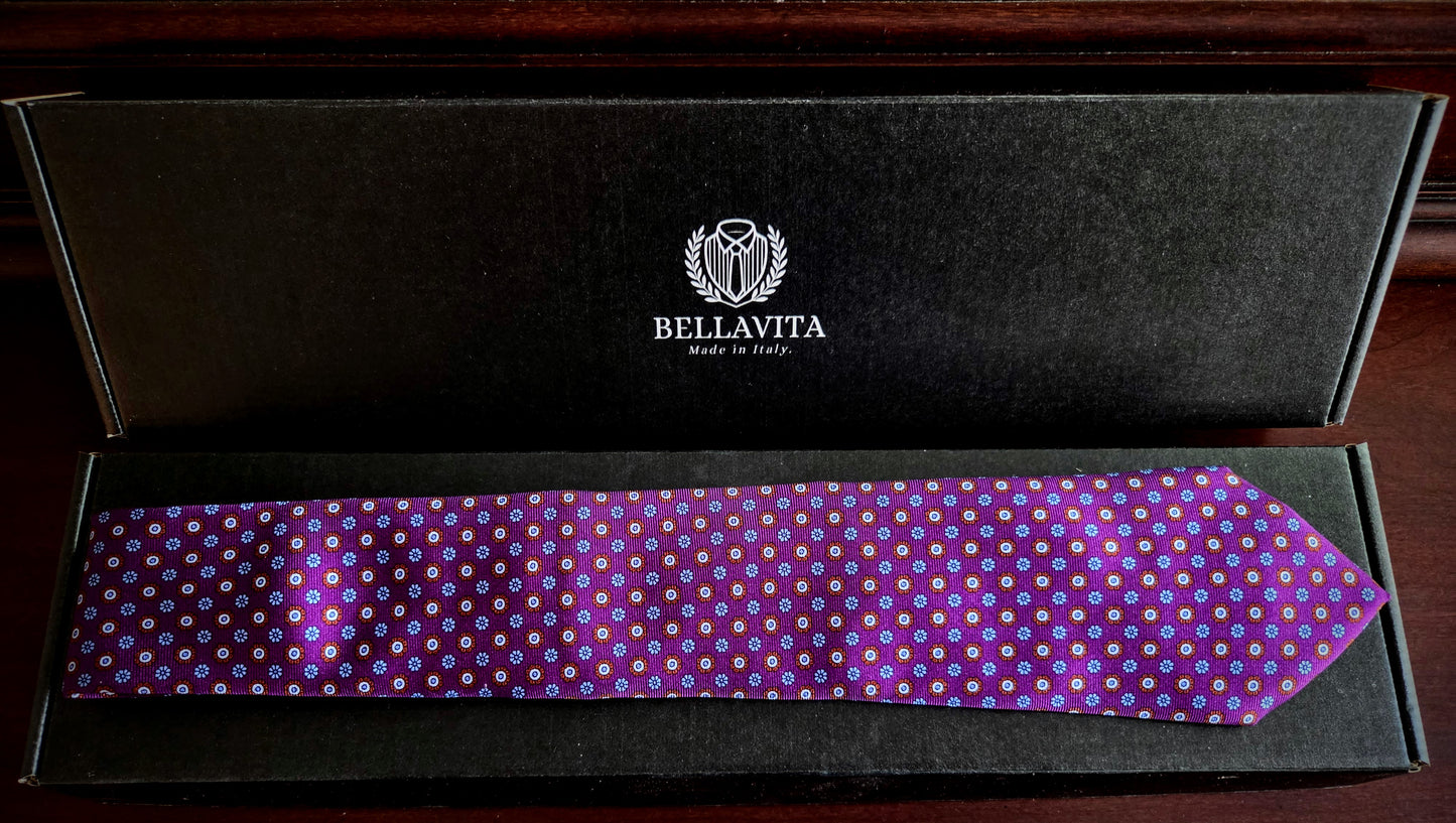 A purple necktie with a white and blue polka dot pattern, presented inside an open black Bellavita gift box on a dark wooden surface.