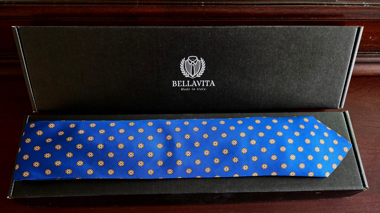 A blue silk necktie with a repeating gold geometric pattern, presented inside its black branded Bellavita gift box on a dark wooden surface.