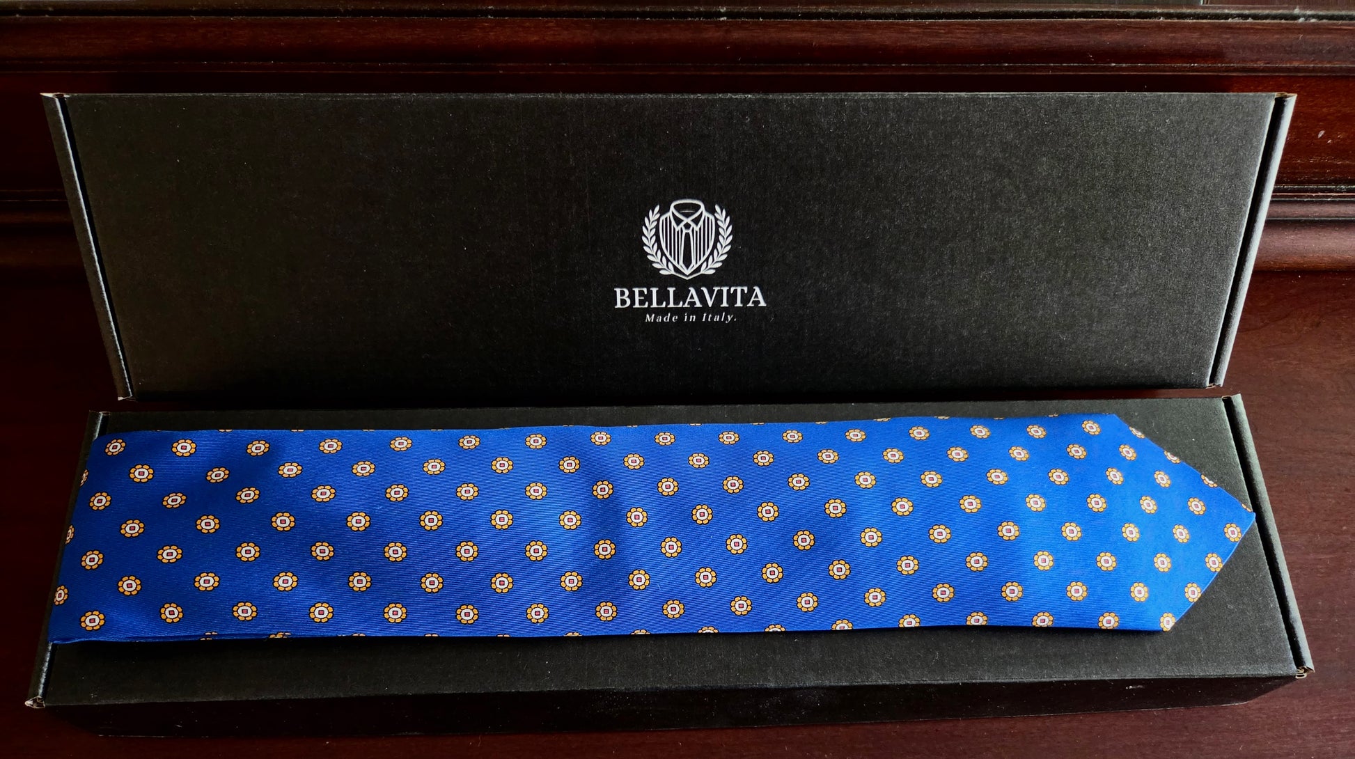 A blue silk necktie with a repeating gold geometric pattern, presented inside its black branded Bellavita gift box on a dark wooden surface.