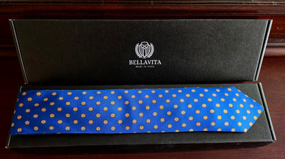 A blue silk necktie with a repeating gold geometric pattern, presented inside its black branded Bellavita gift box on a dark wooden surface.