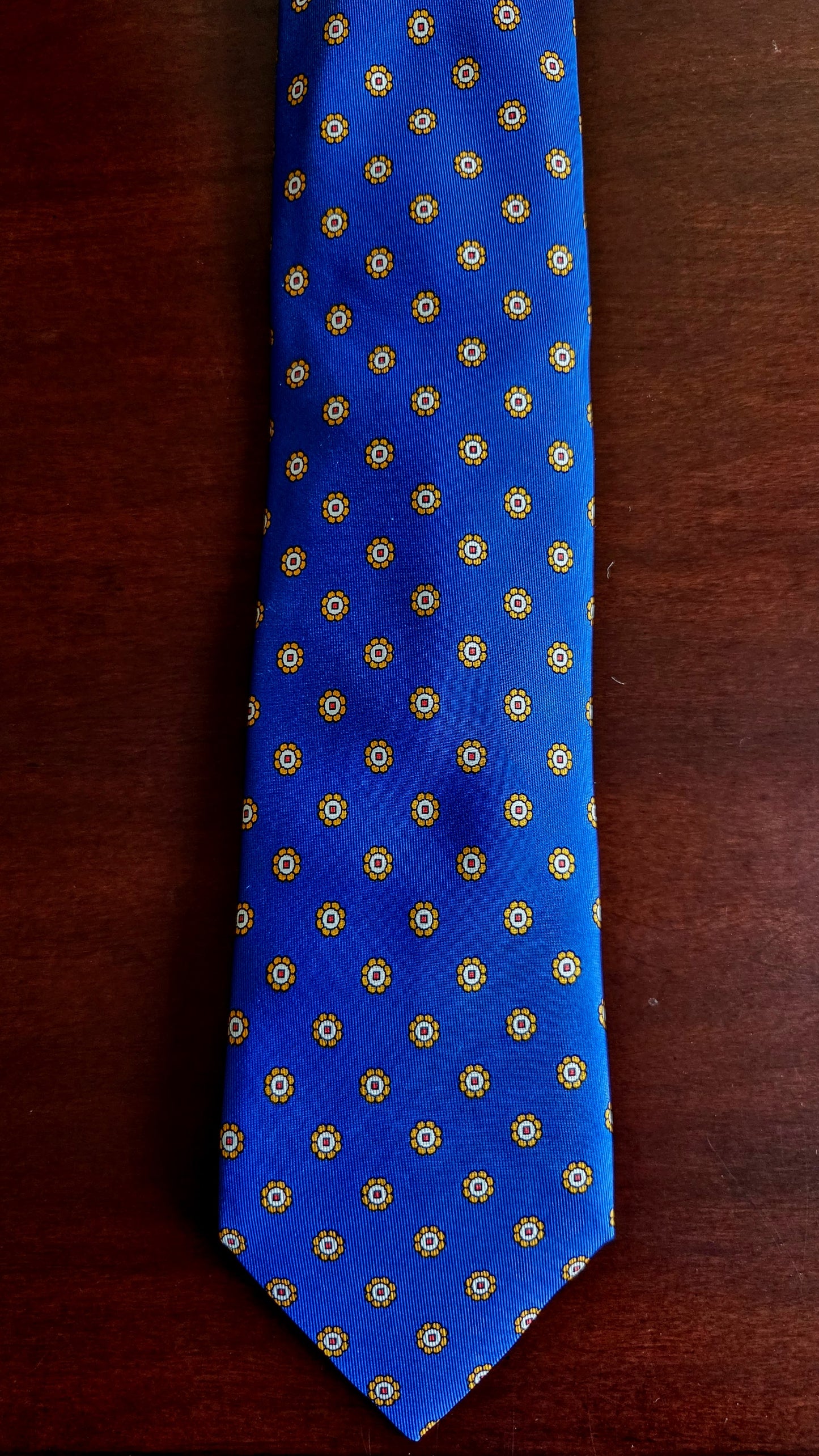 A blue necktie with a repeating gold geometric pattern, laid flat on a dark brown wooden surface.