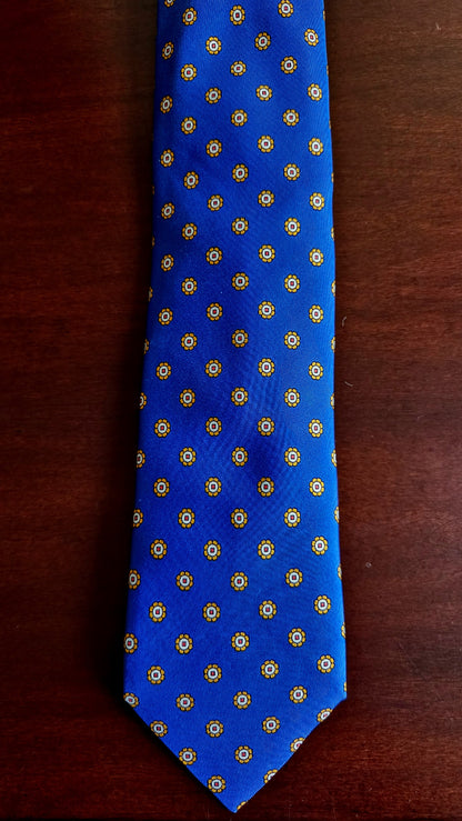 A blue necktie with a repeating gold geometric pattern, laid flat on a dark brown wooden surface.