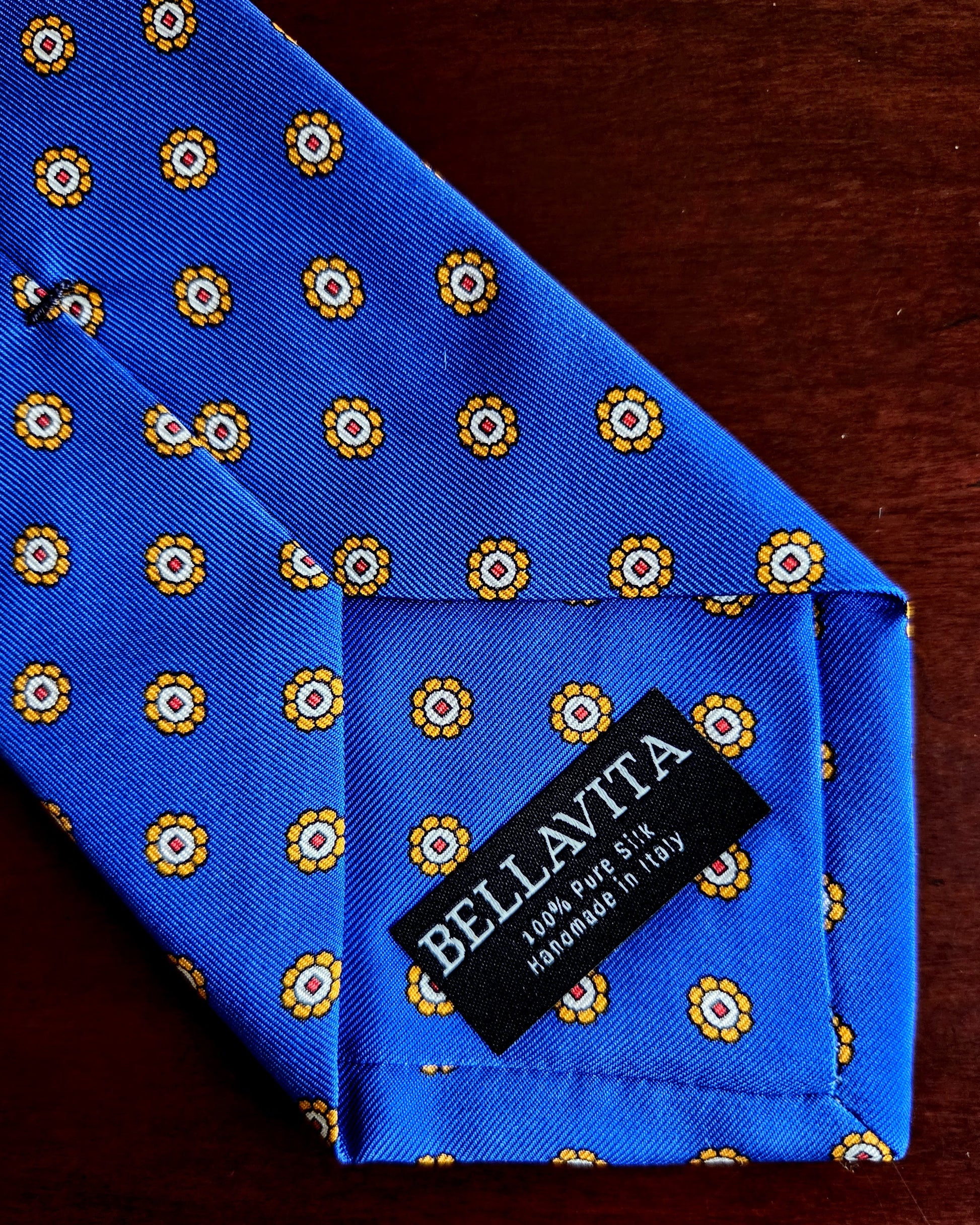 A close-up of a blue silk necktie with a repeating yellow and white floral pattern, featuring a black fabric brand label that reads 'BELLAVITA'.