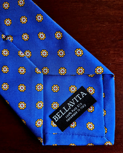 A close-up of a blue silk necktie with a repeating yellow and white floral pattern, featuring a black fabric brand label that reads 'BELLAVITA'.