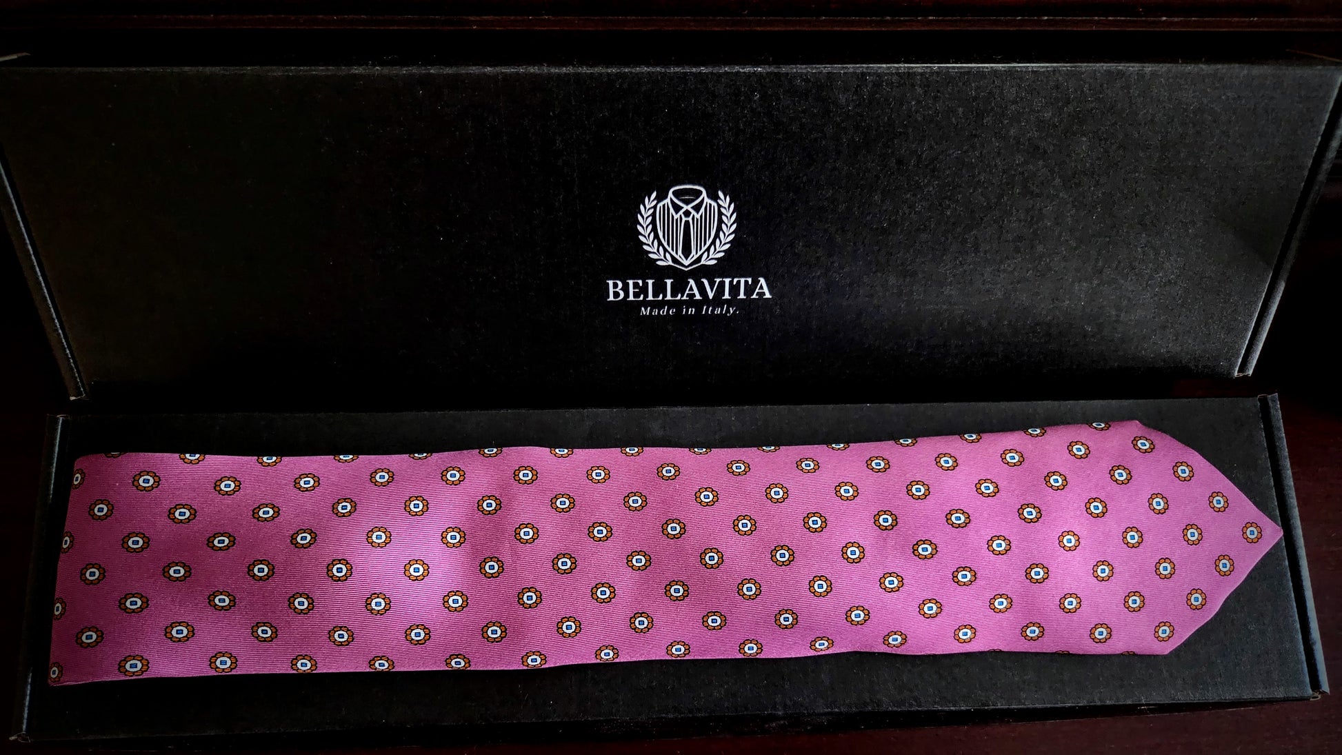 A pink silk necktie with a repeating circular pattern, displayed inside an open black Bellavita gift box.