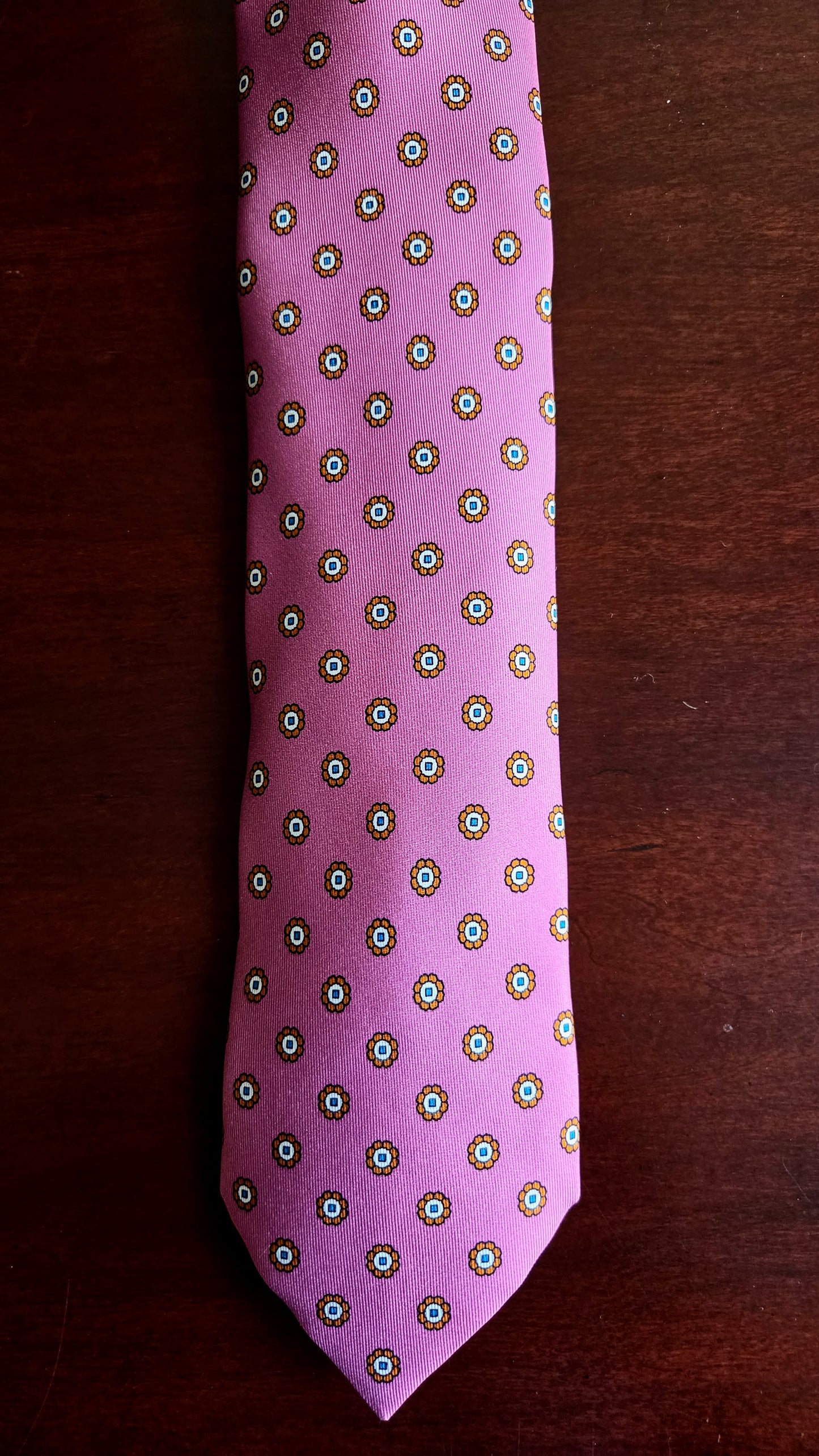 A pink necktie with a repeating geometric medallion pattern in green, white, and gold, laid flat on a dark brown wooden surface.