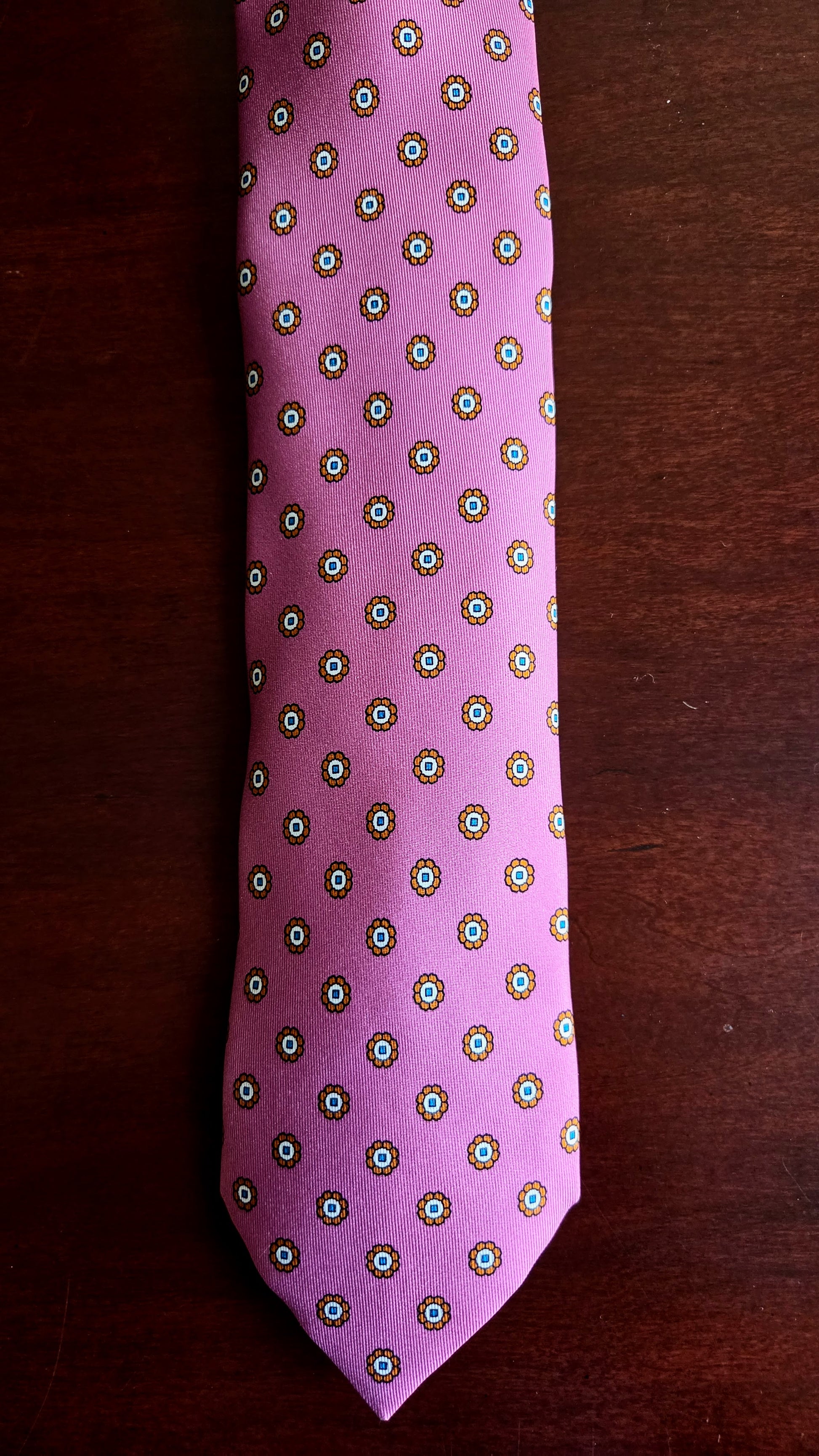 A pink necktie with a repeating geometric medallion pattern in green, white, and gold, laid flat on a dark brown wooden surface.