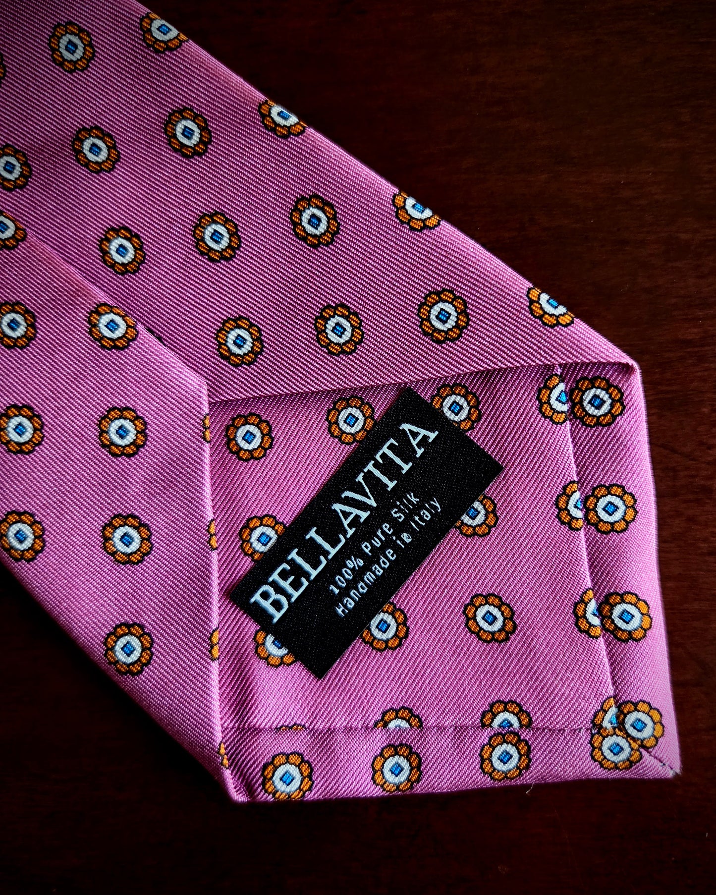 A pink silk necktie with a repeating circular floral pattern in yellow, brown, and blue, featuring a black fabric brand label that reads 'BELLAVITA' and '100% Pure Silk Handmade in Italy.'