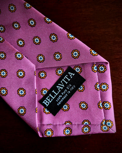 A pink silk necktie with a repeating circular floral pattern in yellow, brown, and blue, featuring a black fabric brand label that reads 'BELLAVITA' and '100% Pure Silk Handmade in Italy.'