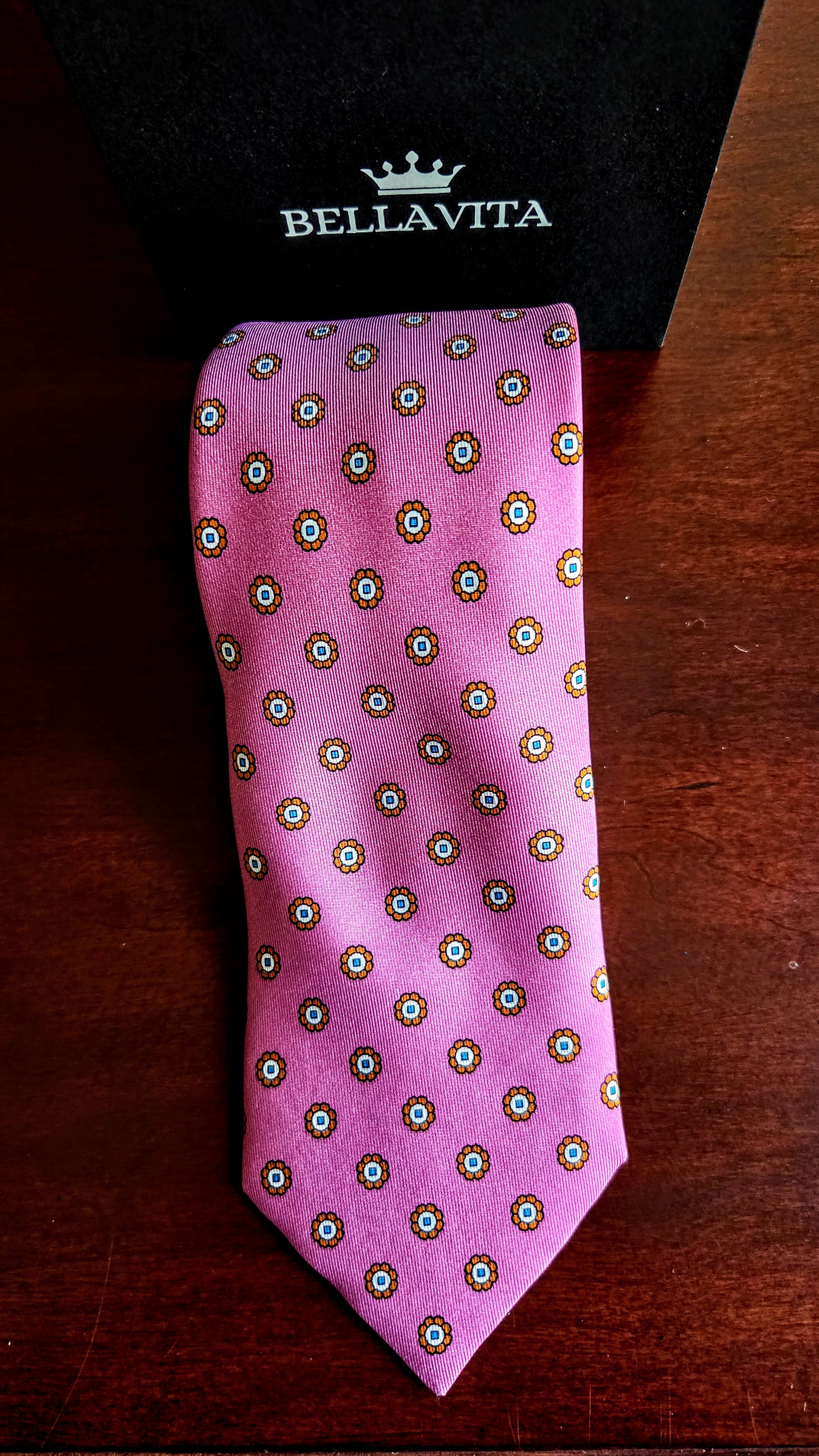 A pink silk necktie with a repeating circular medallion pattern in green, white, and gold, displayed next to its black Bellavita gift box on a dark wooden surface.