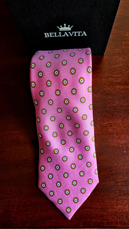 A pink silk necktie with a repeating circular medallion pattern in green, white, and gold, displayed next to its black Bellavita gift box on a dark wooden surface.
