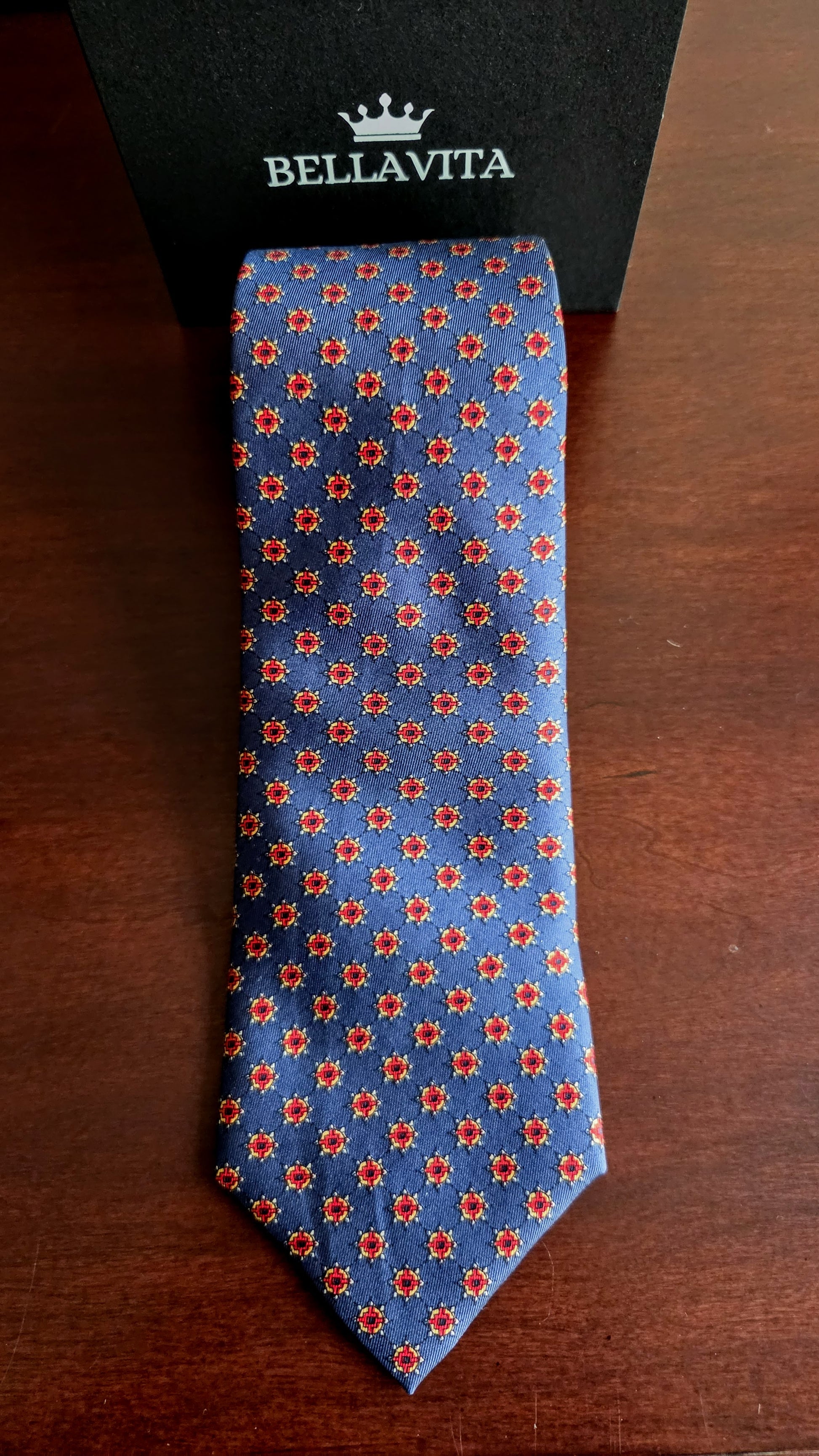 A blue silk necktie with a repeating red and white floral pattern, displayed vertically on a dark wooden surface next to its black Bellavita gift box.