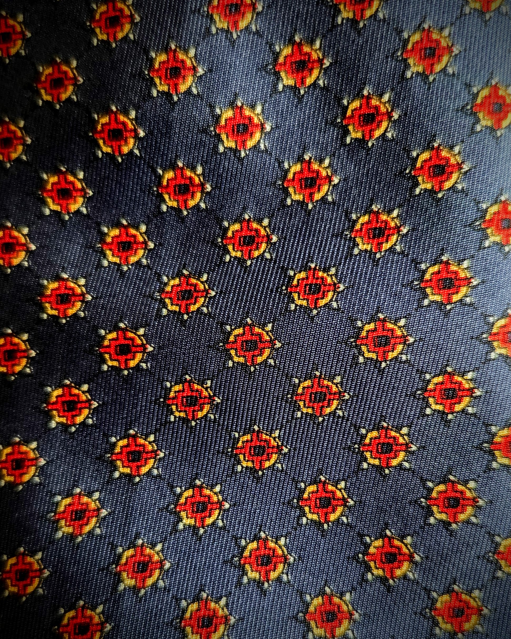 A close-up of a woven necktie fabric featuring a repeating geometric medallion pattern in red, yellow, and black on a dark charcoal gray background.