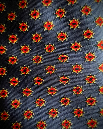A close-up of a woven necktie fabric featuring a repeating geometric medallion pattern in red, yellow, and black on a dark charcoal gray background.
