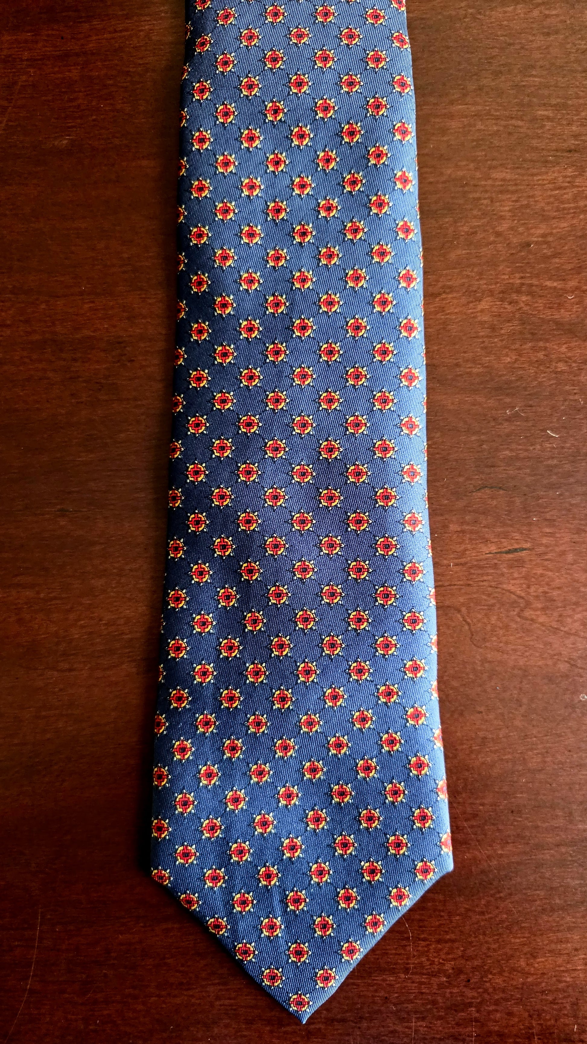 A blue necktie with a repeating red and orange floral pattern, laid flat on a dark brown wooden surface.