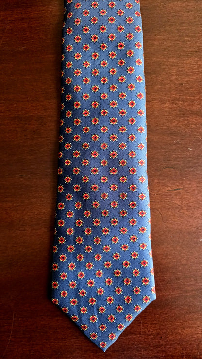 A blue necktie with a repeating red and orange floral pattern, laid flat on a dark brown wooden surface.